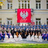 Zu sehen sind Tänzer und Musiker des Künstlerensembles in Uniform und Kostüm vor einem Gebäude, vom Balkon hängt der polnische Wappenadler